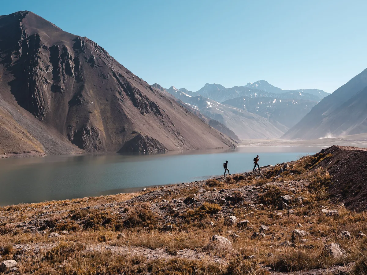 Two hikers walking along the shore of the turquoise Embalse El Yeso reservoir beneath bare Andean peaks